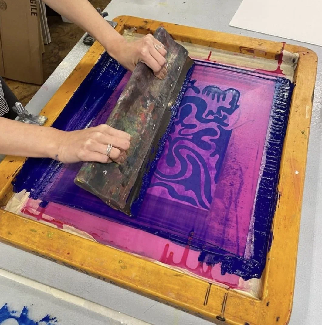 A person pulls a squeegee across a wooden-framed silk screen, pressing vibrant blue ink over a pink mesh screen with an abstract design. The screen shows ink buildup along the edges from repeated use.