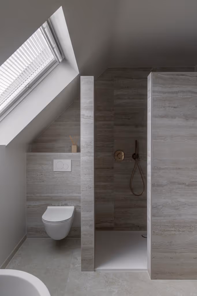 Foto van een moderne badkamer met wit toilet, goude kranen, tegels uit natuursteen en vernieuwde velux.