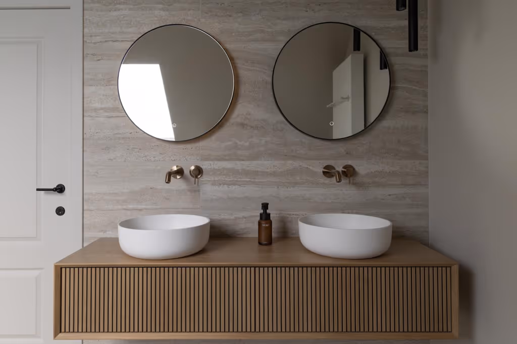 Moderne badkamerrenovatie met witte waskommen, goude kranen, zwarte spiegels en een badkamermeubel uit hout. 