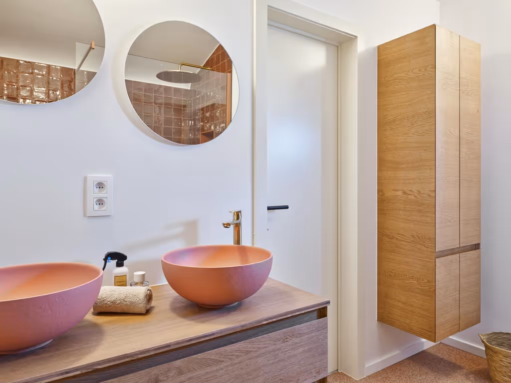 Stijlvolle badkamerrenovatie met houten meubel, roze waskommen, ronde spiegels en warme materialen.