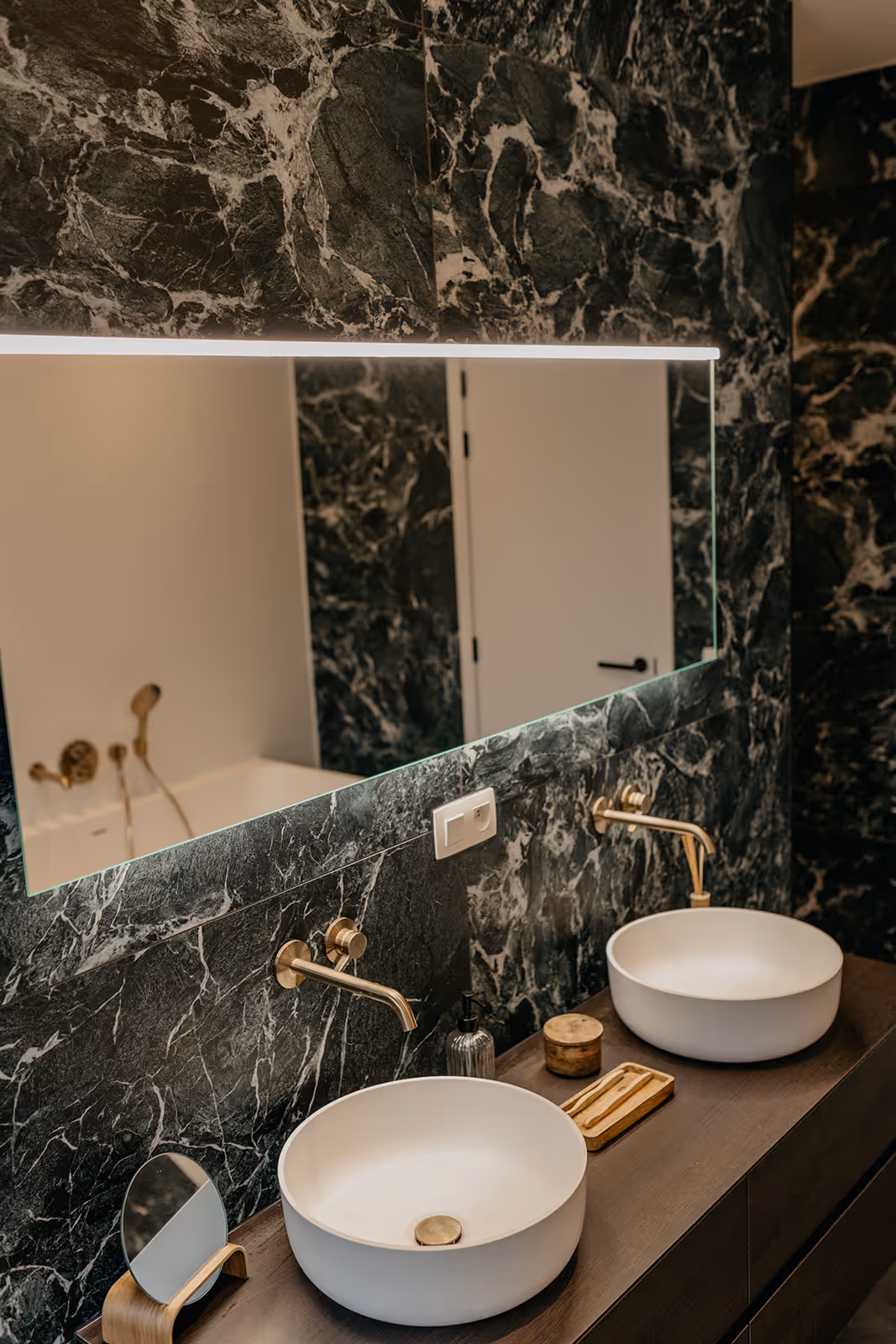 Badkamer na totaalrenovatie met wandtegels uit natuursteen, verlichte spiegel en witte waskommen.