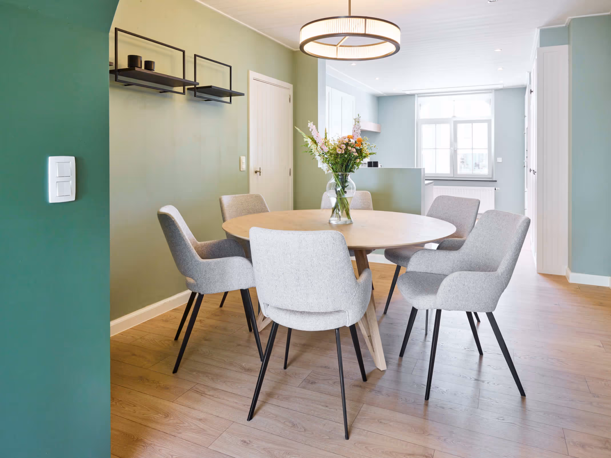 Eetruimte na de totaalrenovatie met groene accentmuur, ronde tafel, houten vloer, en keuken op de achtergrond.