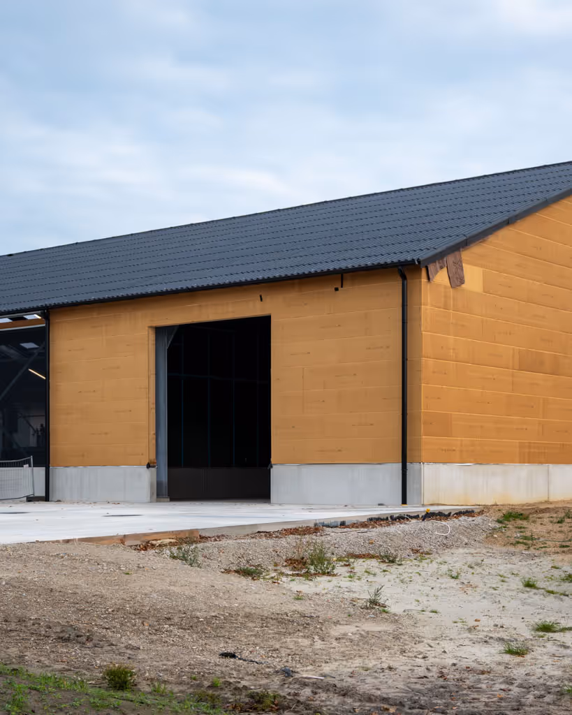 Voorgevel van een nieuwbouwhal met de houtskeletwand, donkere golfplaten en betonnen plint.