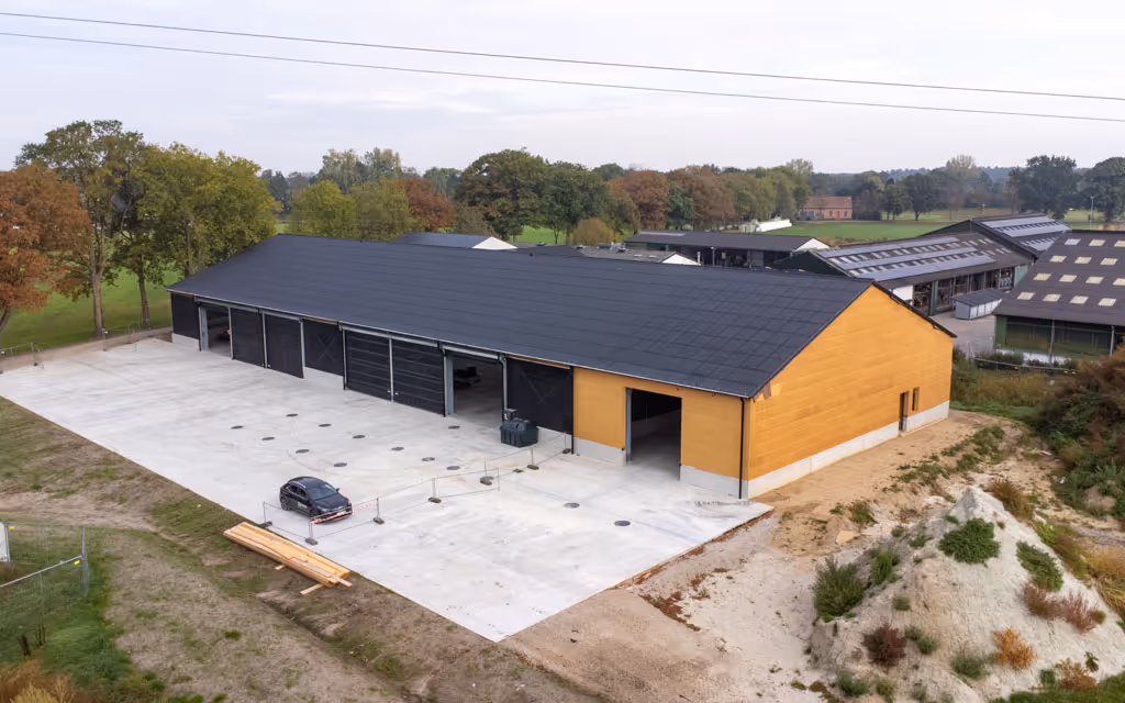 Luchtfoto van een nieuwe hal met wanden uit houtskelet en donkere dakbedekking op het hellende dak.