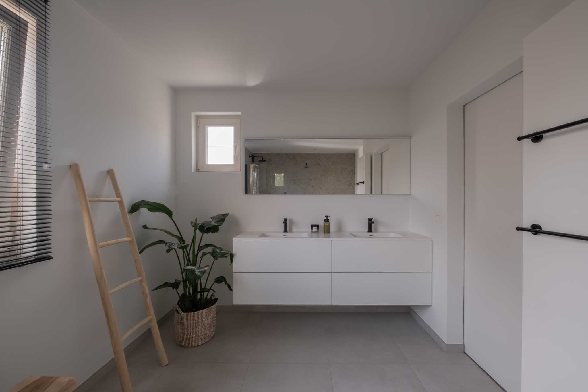 Moderne minimalistische badkamerrenovatie met wit badkamermeubel, dubbele wastafel, zwarte kranen, grote spiegel en grijze tegelvloer.