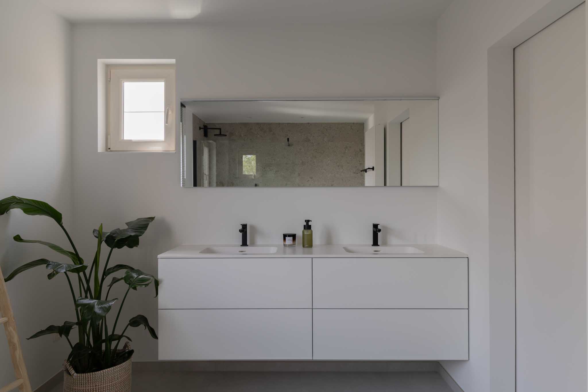 Moderne minimalistische badkamerrenovatie met wit badkamermeubel, dubbele wastafel, zwarte kranen, grote spiegel en grijze tegelvloer.