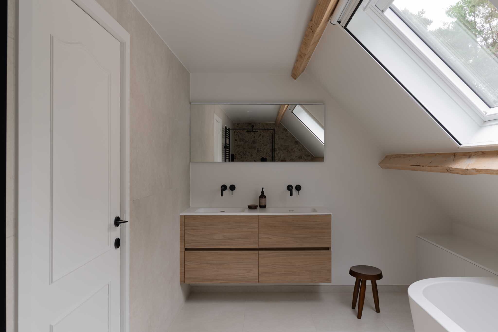 Moderne badkamerrenovatie met natuurlijke tinten, zwarte kranen, witte maatkasten en zichtbare houten gordingen voor luxe en warmte.