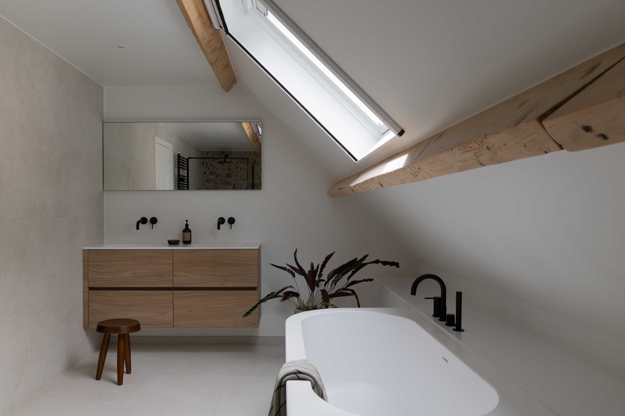 Moderne badkamerrenovatie met natuurlijke tinten, zwarte kranen, witte maatkasten en zichtbare houten gordingen voor luxe en warmte.