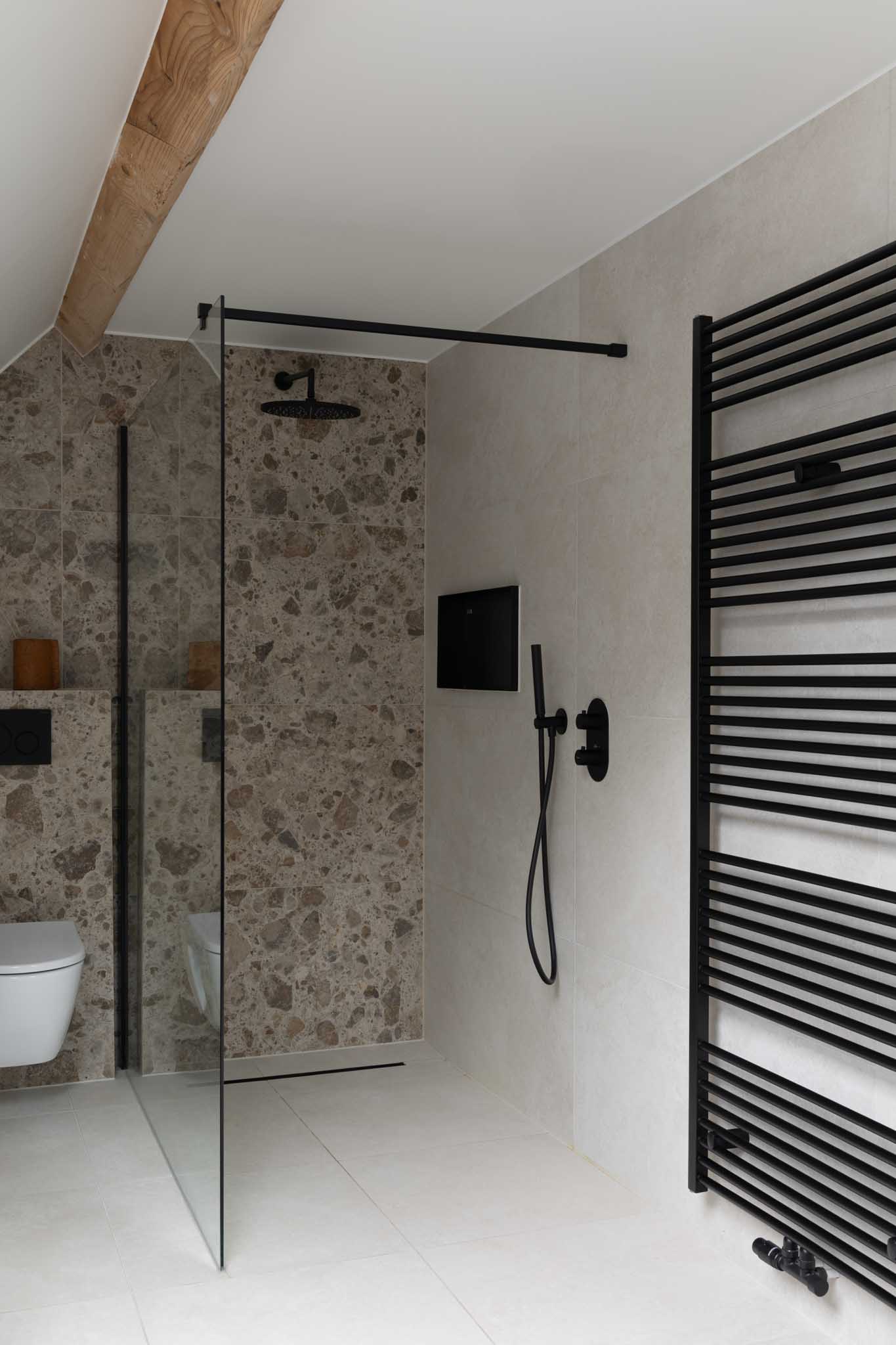 Moderne badkamerrenovatie met zwarte radiator, inloopdouche met zwart kraanwerk, natuurstenen tegels en wit toilet;