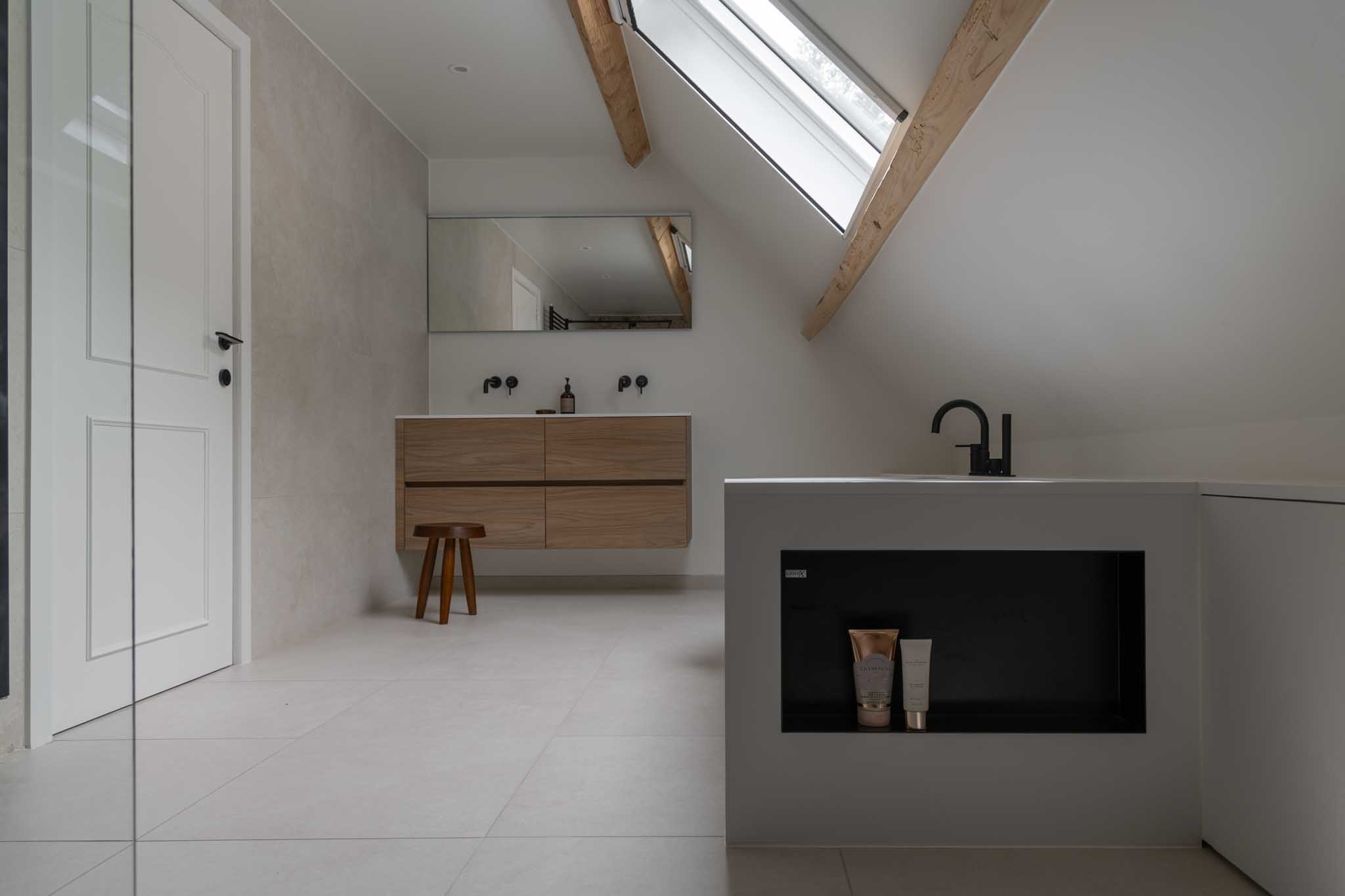 Moderne badkamerrenovatie met inbouwnis achter het back-to-wall bad, beige tegels, houten badkamermeubel en grote spiegel.