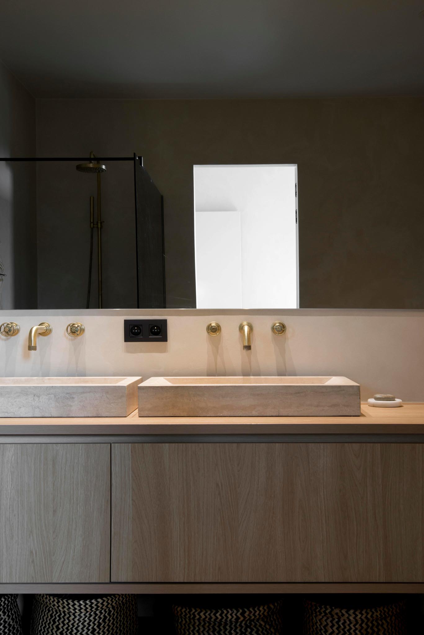 Moderne badkamerrenovatie met dubbele spoelbak op houten badkamermeubel, gouden kranen en grote spiegel.