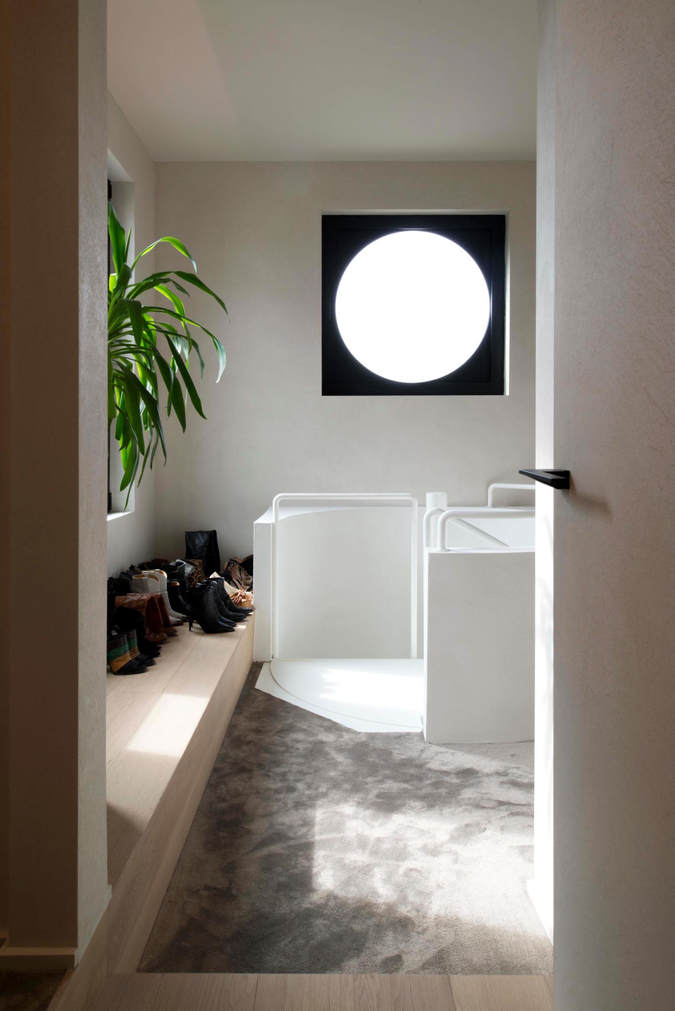 Nachthal na totaalrenovatie met ronde trap, lichte kleuren, houten zitbank met plaats voor schoenen en rond antraciet raam.