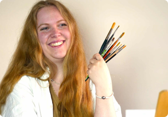 Smiling young woman with long red hair holding multiple paintbrushes in her hand.