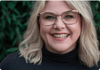 Smiling blonde woman with glasses wearing a black turtleneck in front of greenery.