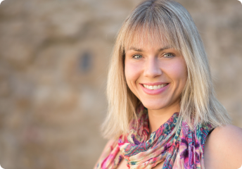 Smiling blonde woman with a colorful scarf standing in front of a blurred stone wall.
