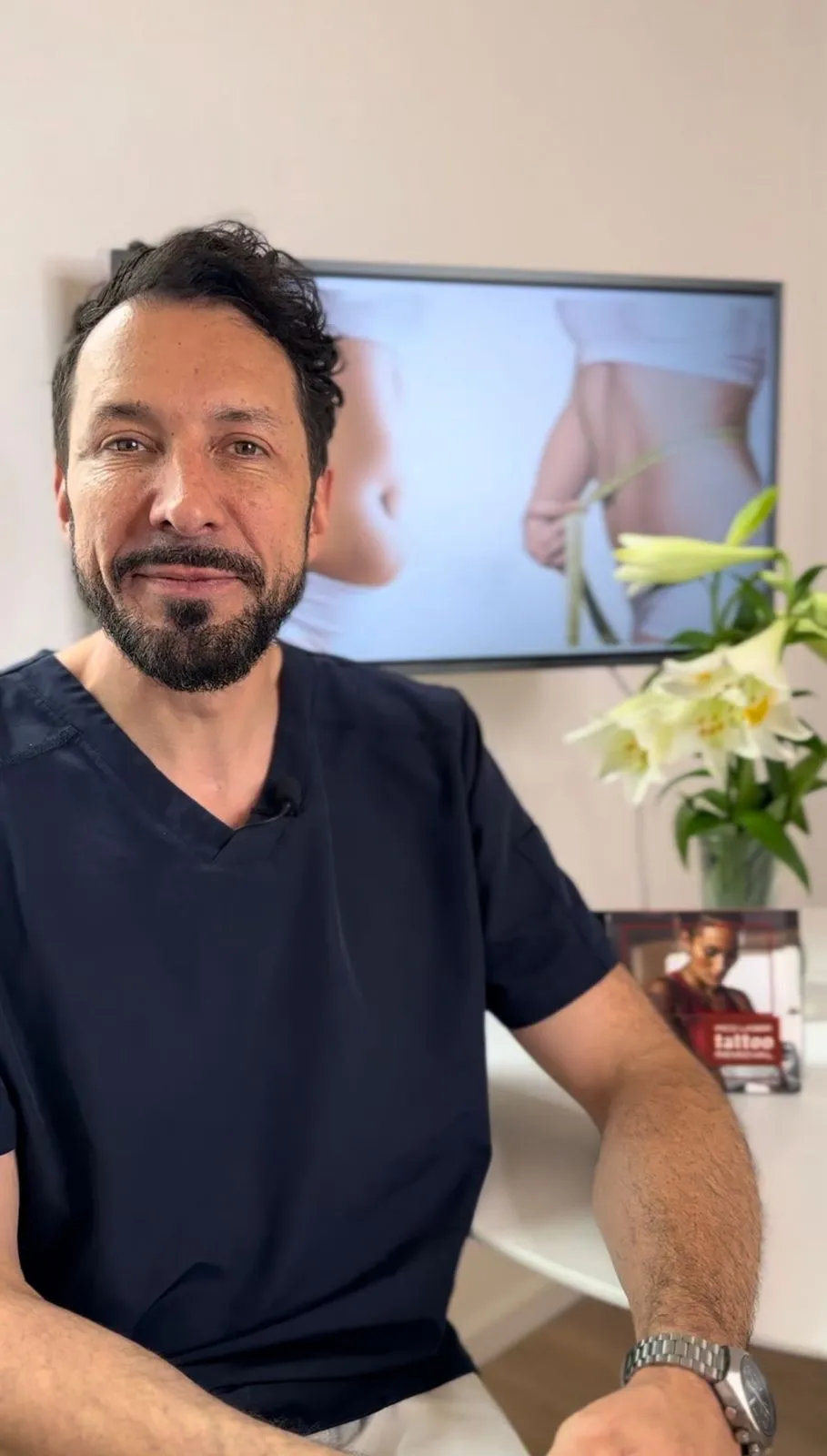 Man with dark hair and beard wearing navy scrubs sitting indoors with a TV screen showing a close-up of a person's waist being measured and a vase of white lilies in the background.