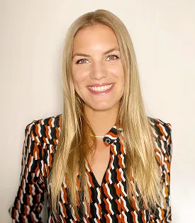 Lächelnde blonde Frau mit langem Haar, die ein schwarz-orange-weiß gemustertes Hemd trägt.