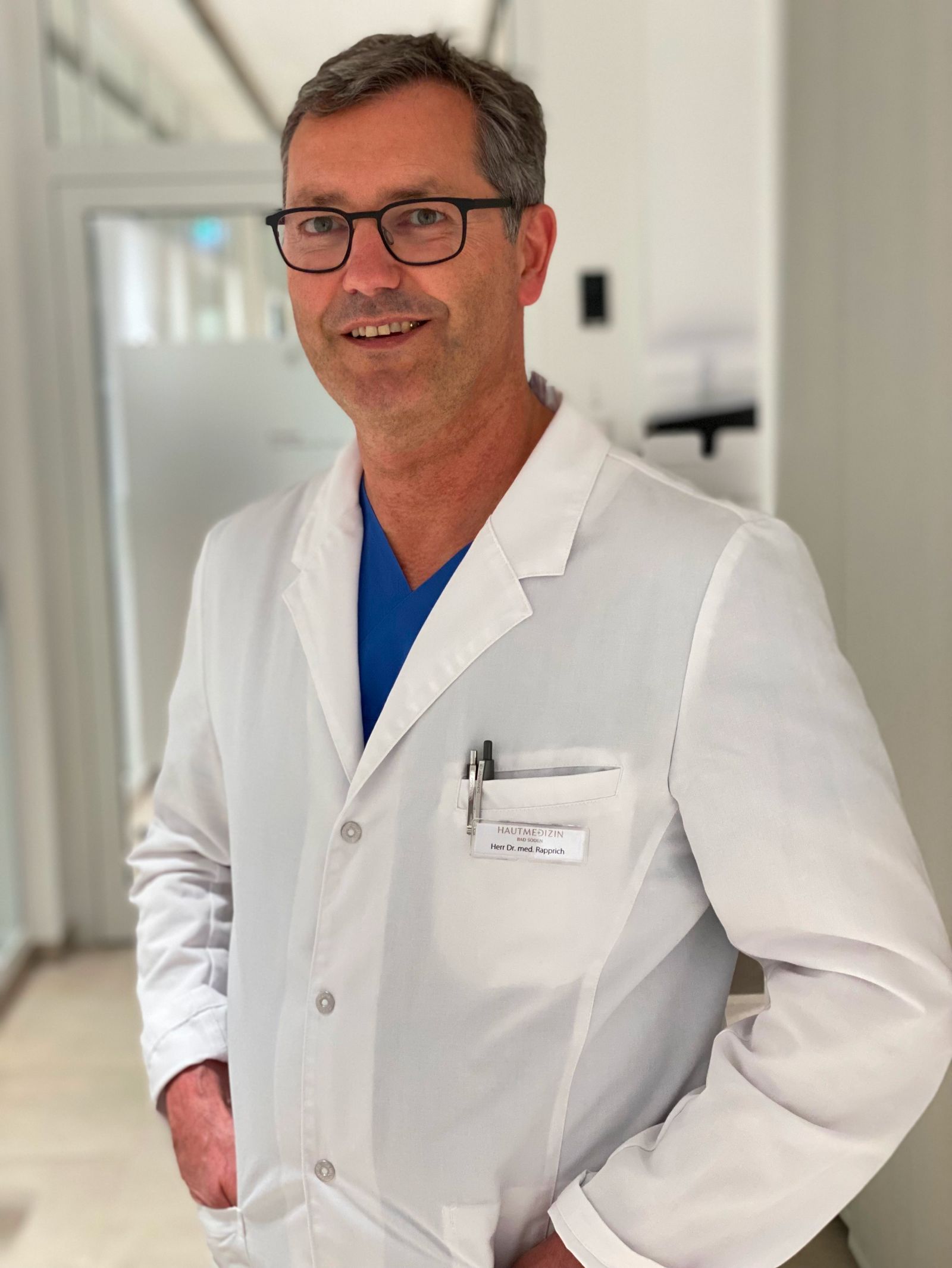 Männlicher Arzt in weißem Kittel mit Namensschild und Brille in einem hellen Flur.