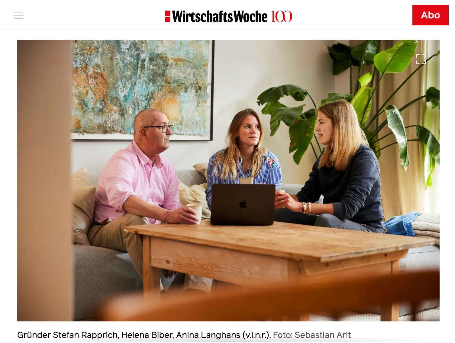 Drei Personen sitzen auf einem Sofa um einen Holztisch mit einem Laptop und halten Tassen in den Händen, mit Pflanzen und einem abstrakten Gemälde im Hintergrund.