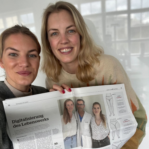 Zwei lächelnde Frauen, eine in grauem Pullover und eine in hellem Pullover, halten eine Zeitung mit dem Titel 'Digitalisierung des Lebenswerks' mit Fotos von drei Personen in medizinischer Kleidung.