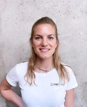 Junge Frau mit blondem Haar trägt ein weißes T-Shirt vor einer Betonwand.