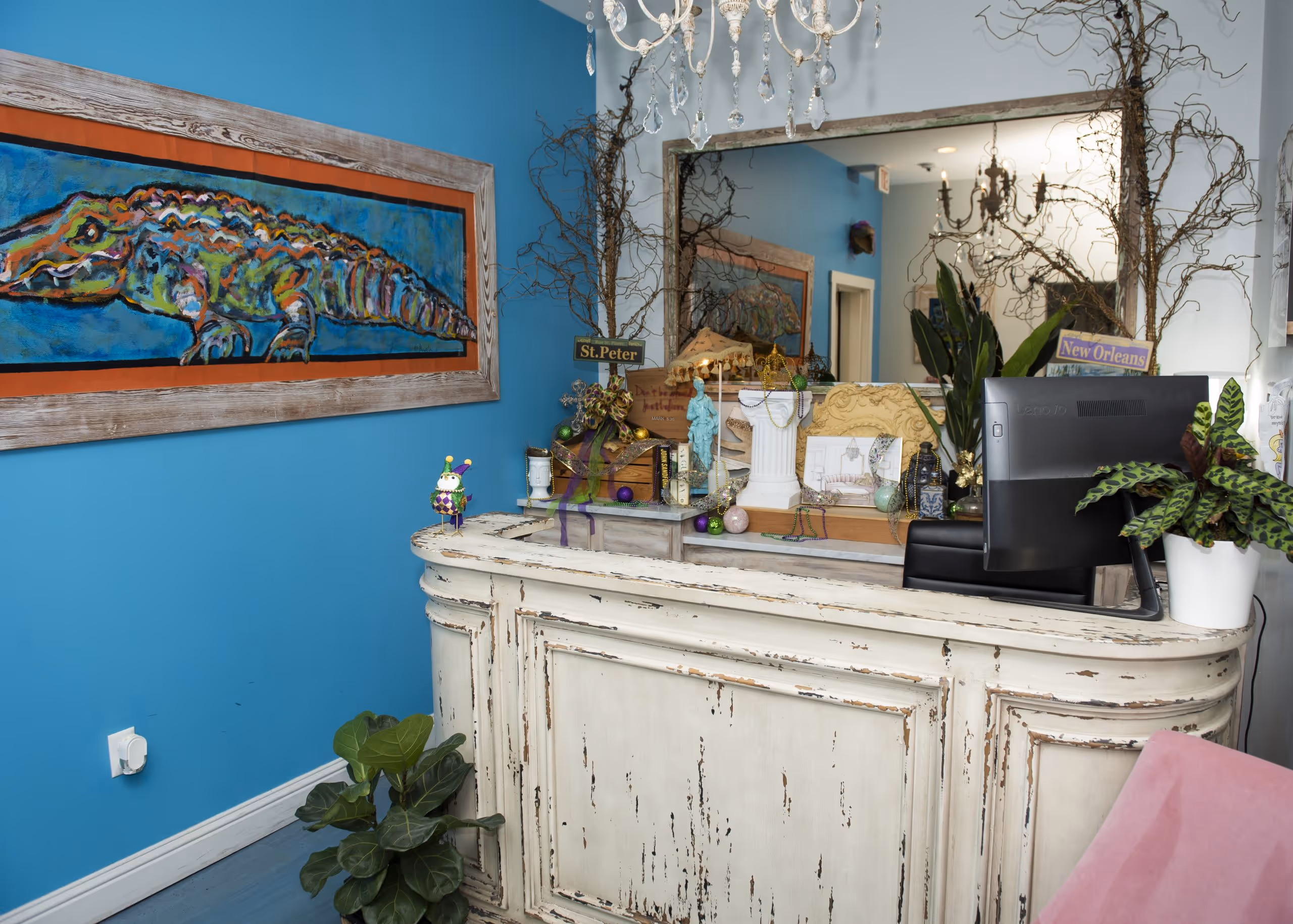 Vintage-style white reception desk with decorative items, a computer monitor, plants, and a colorful alligator painting on a blue wall.