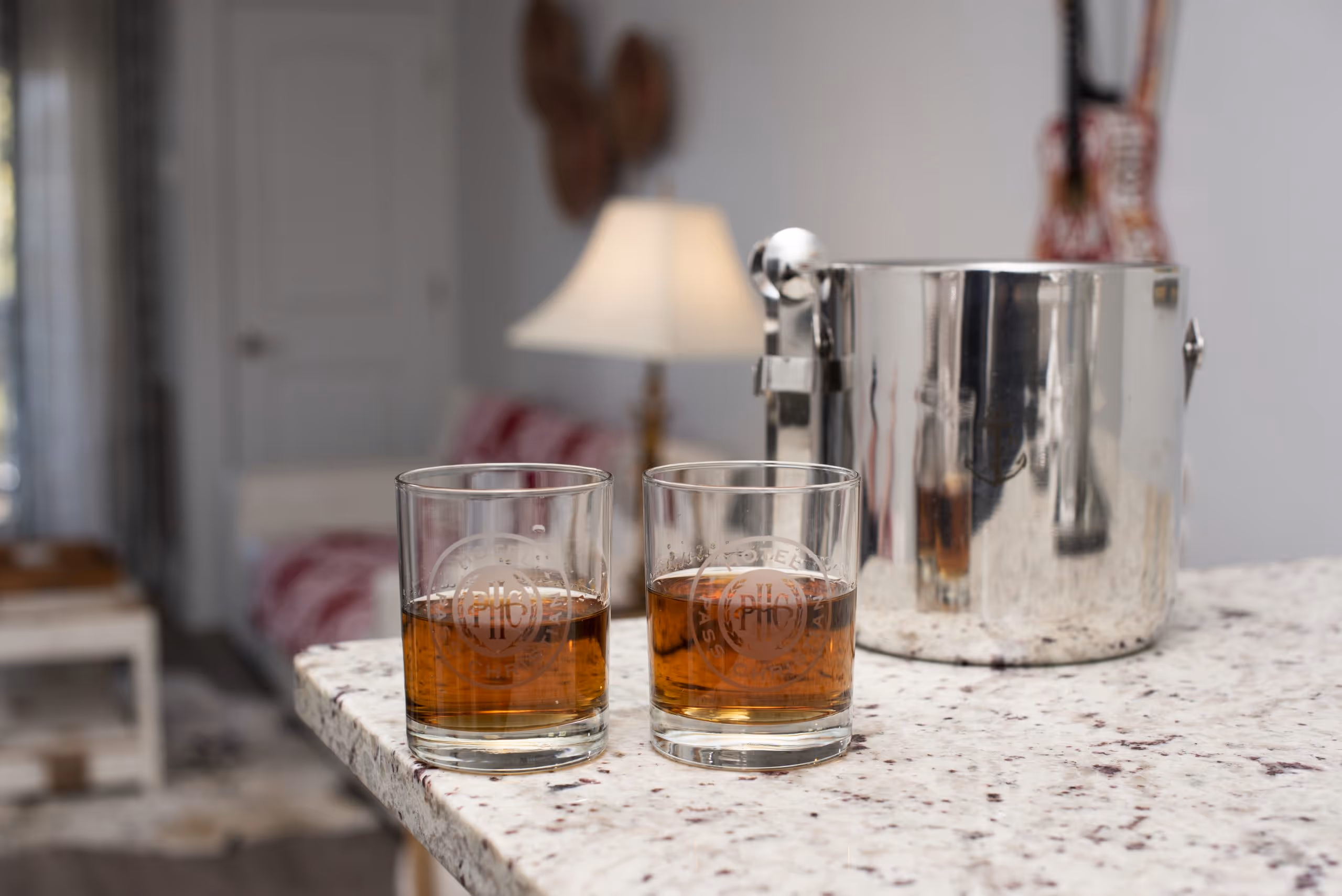 Two glasses with bourbon on a granite countertop next to a metallic ice bucket in a cozy room.