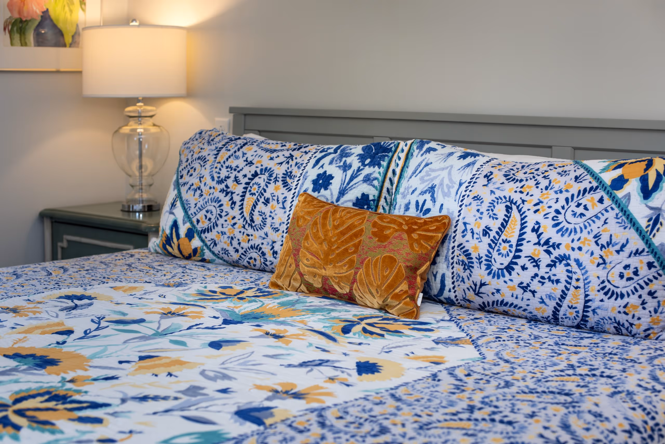 Bed with a blue, yellow, and white floral patterned quilt and pillows, featuring a small orange decorative pillow, next to a green nightstand with a glass lamp.