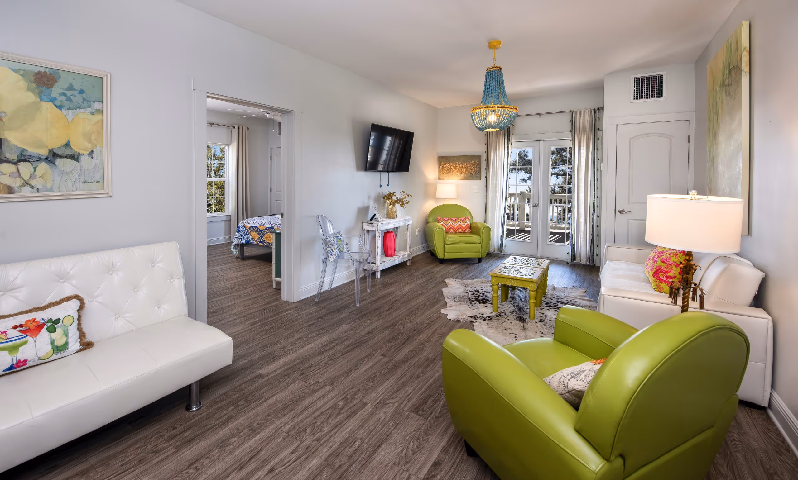 Modern living room with green armchairs, white sofas, wood flooring, colorful cushions, wall-mounted TV, and a doorway leading to a bedroom.