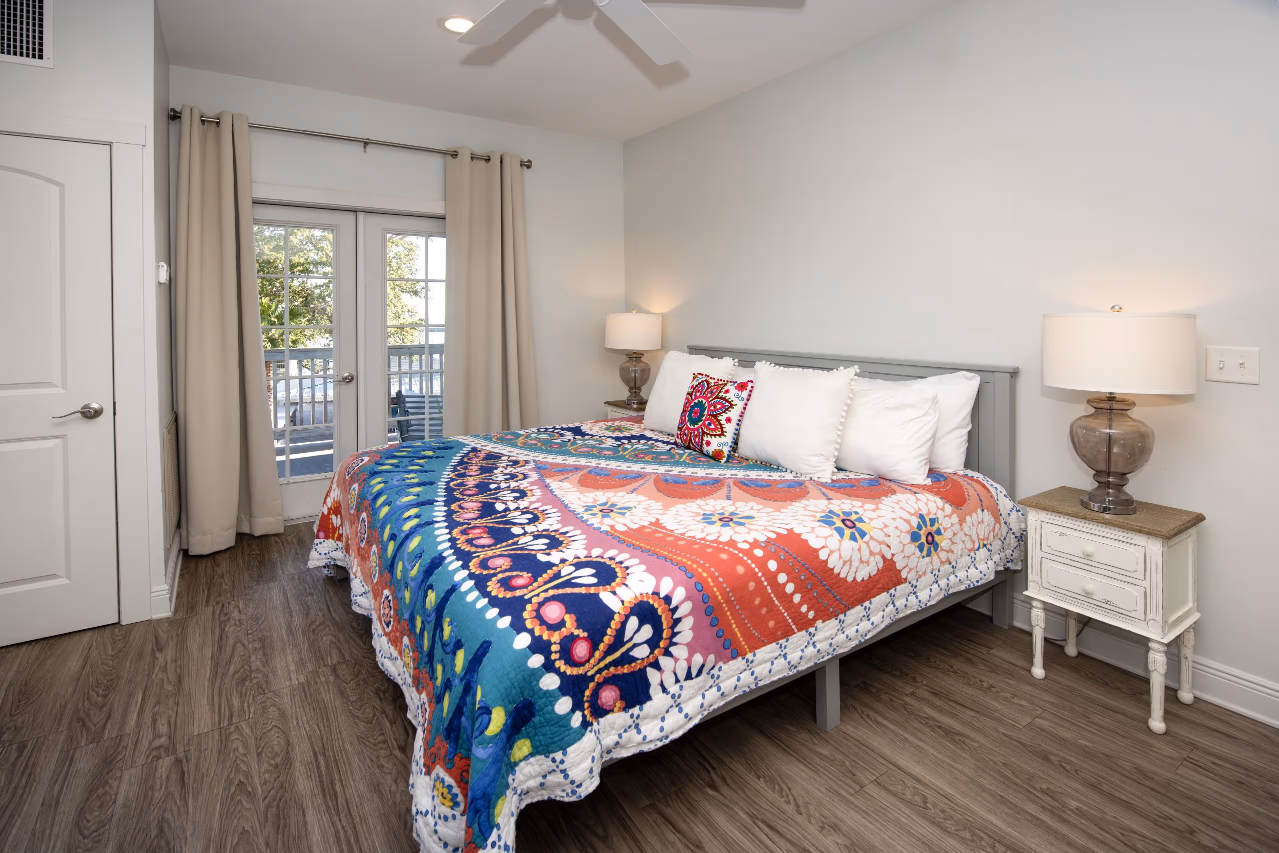 Bedroom with a large bed featuring a colorful patterned quilt and white pillows, two nightstands with lamps, and glass doors with beige curtains.