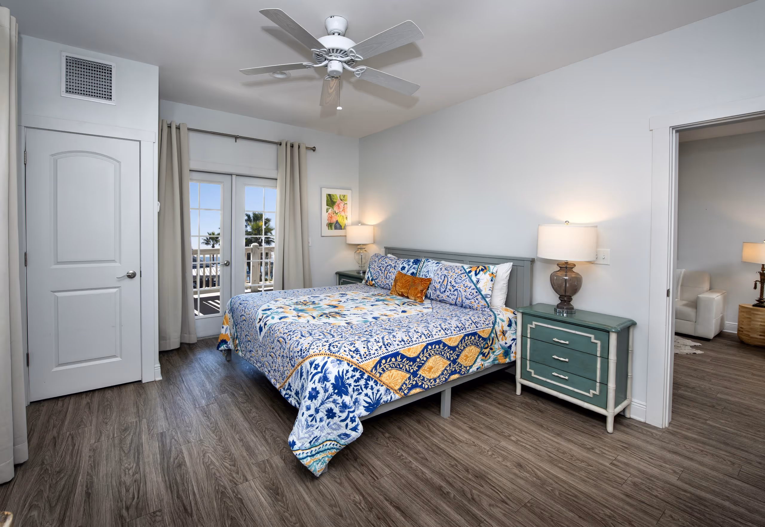 Bright bedroom with large bed featuring a blue, white, and yellow floral quilt, green nightstands with lamps, wood flooring, and glass doors leading to a balcony with palm trees outside.