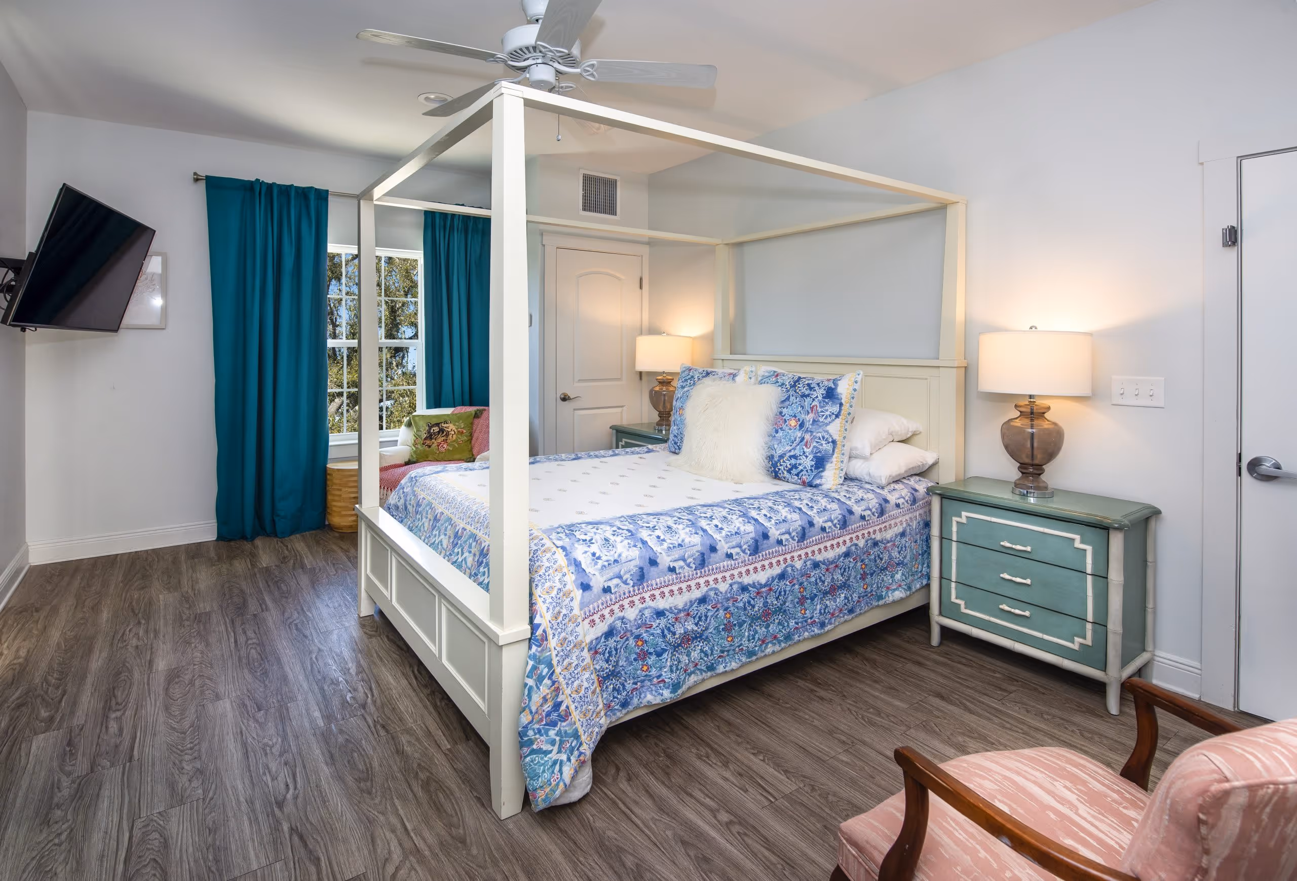 Bright bedroom with a white four-poster bed featuring blue floral bedding, teal nightstand with lamp, teal curtains, wall-mounted TV, and a pink armchair.