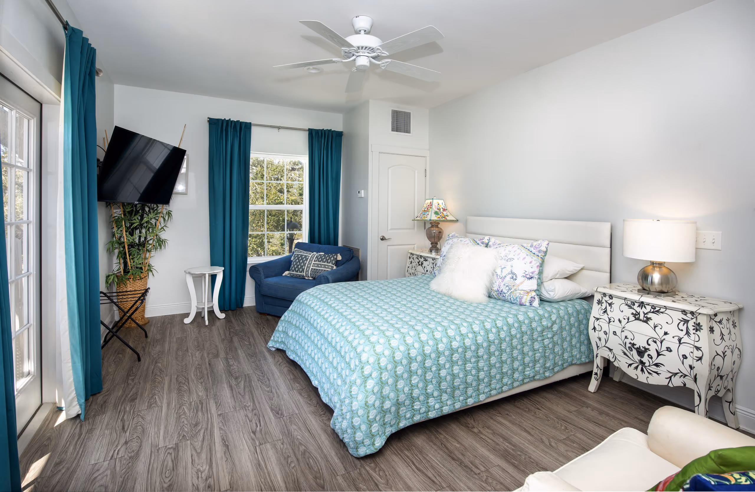 Bright bedroom with a queen bed covered in a blue patterned quilt, teal curtains, a blue armchair by the window, two white nightstands with lamps, and a wall-mounted TV.