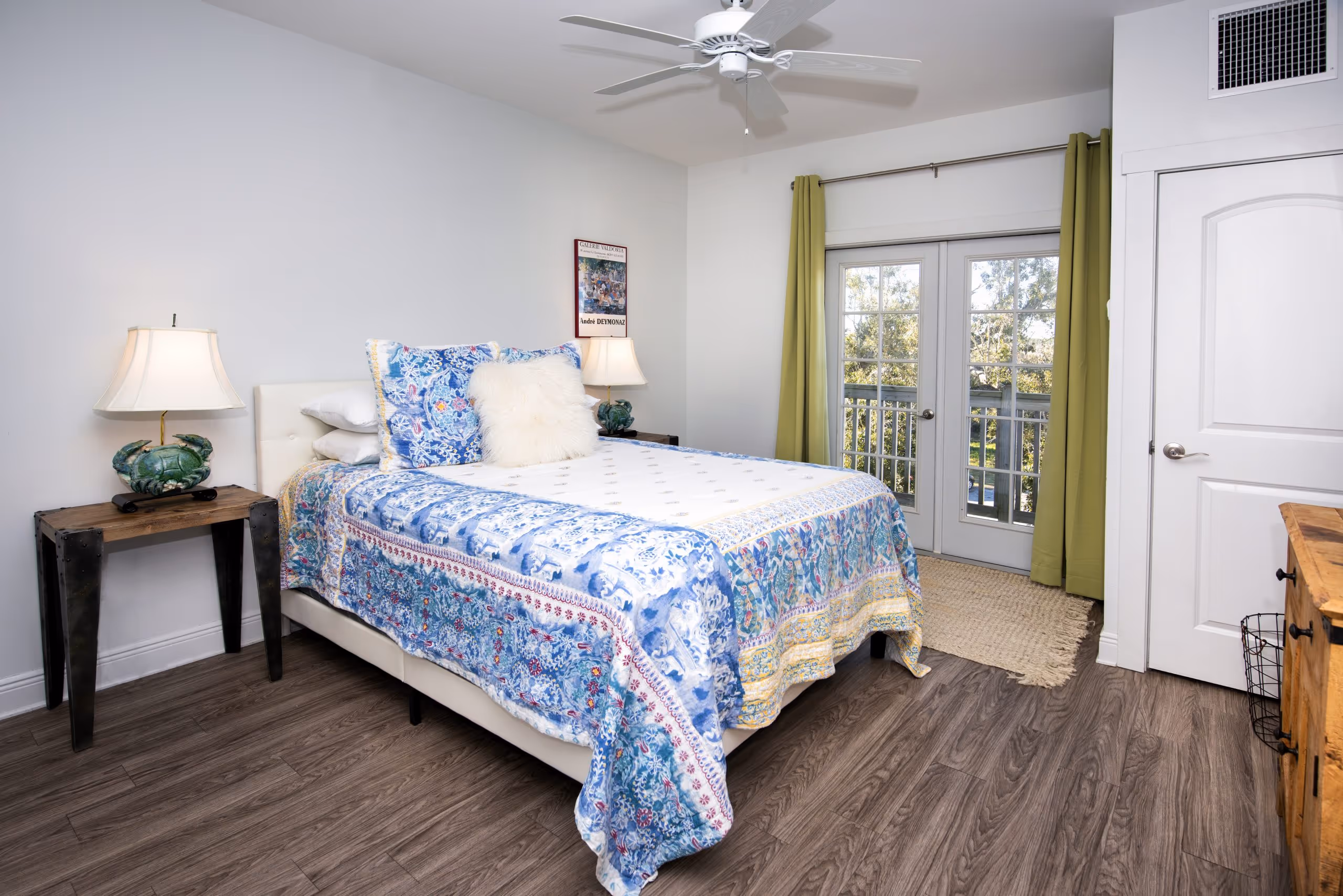 Bedroom with a bed featuring blue and white patterned bedding, two crab-shaped lamps on wooden side tables, green curtains, and glass double doors leading outside.