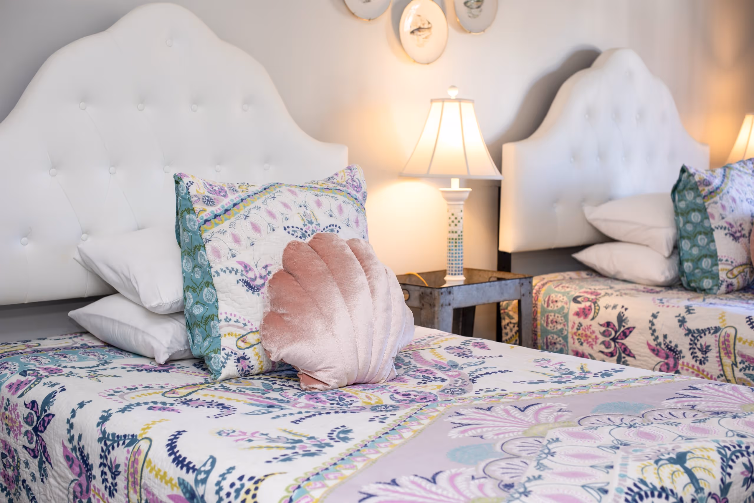 Two beds with white tufted headboards, floral patterned bedding, decorative pillows including a pink shell-shaped pillow, and a lamp on a nightstand between them.