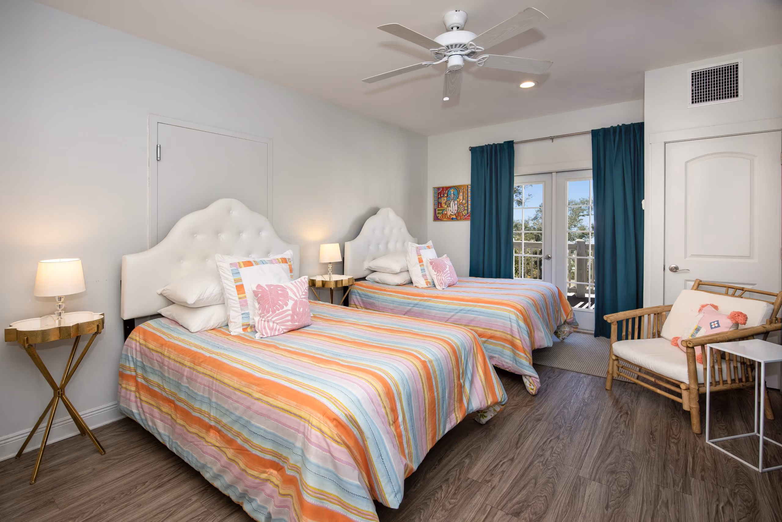 Bedroom with two twin beds featuring white tufted headboards, colorful striped bedspreads, and pink leaf-patterned pillows, lit by bedside lamps.