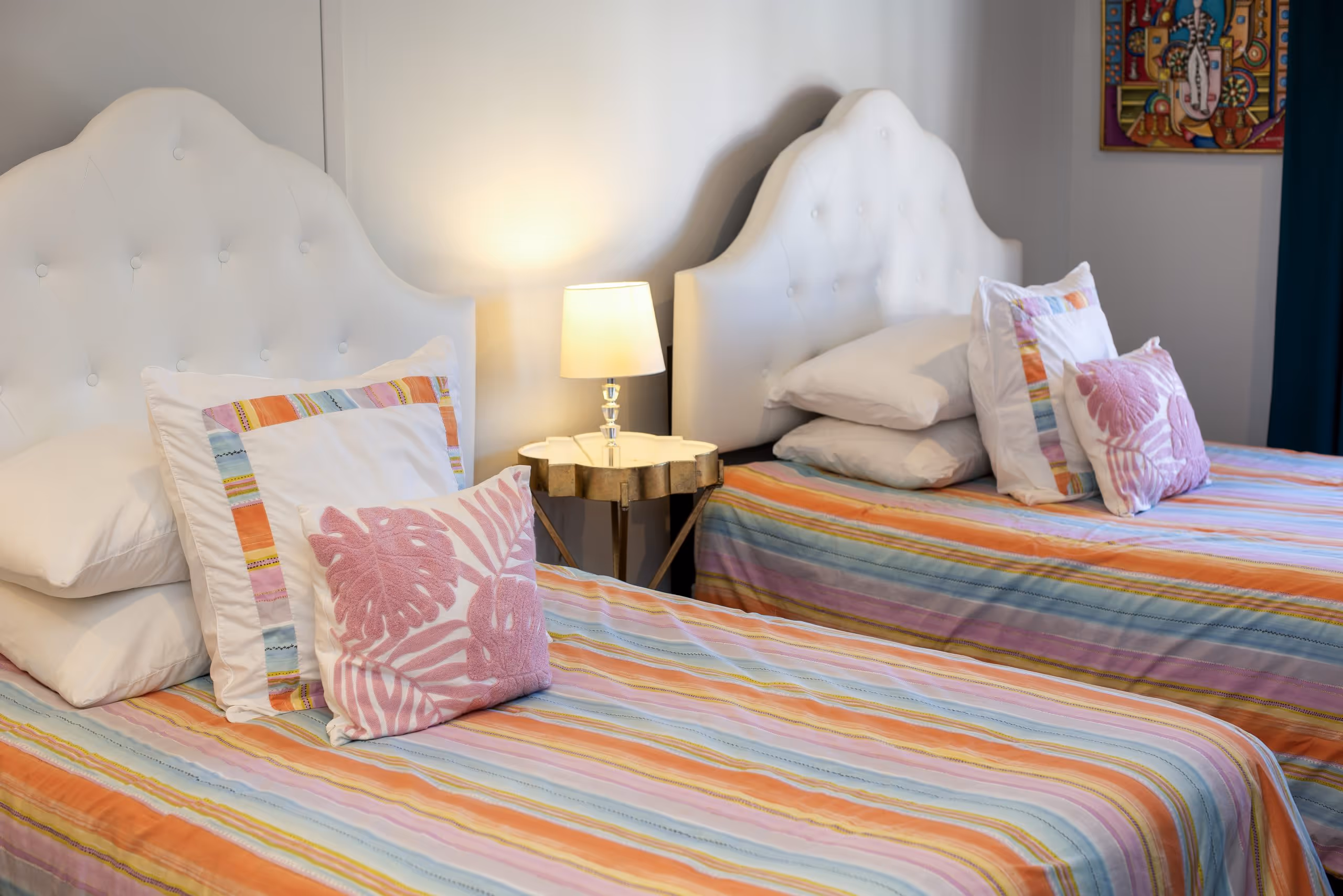 Two twin beds with white tufted headboards, colorful striped bedspreads, and decorative pillows with pink leaf patterns, separated by a small table with a lamp.