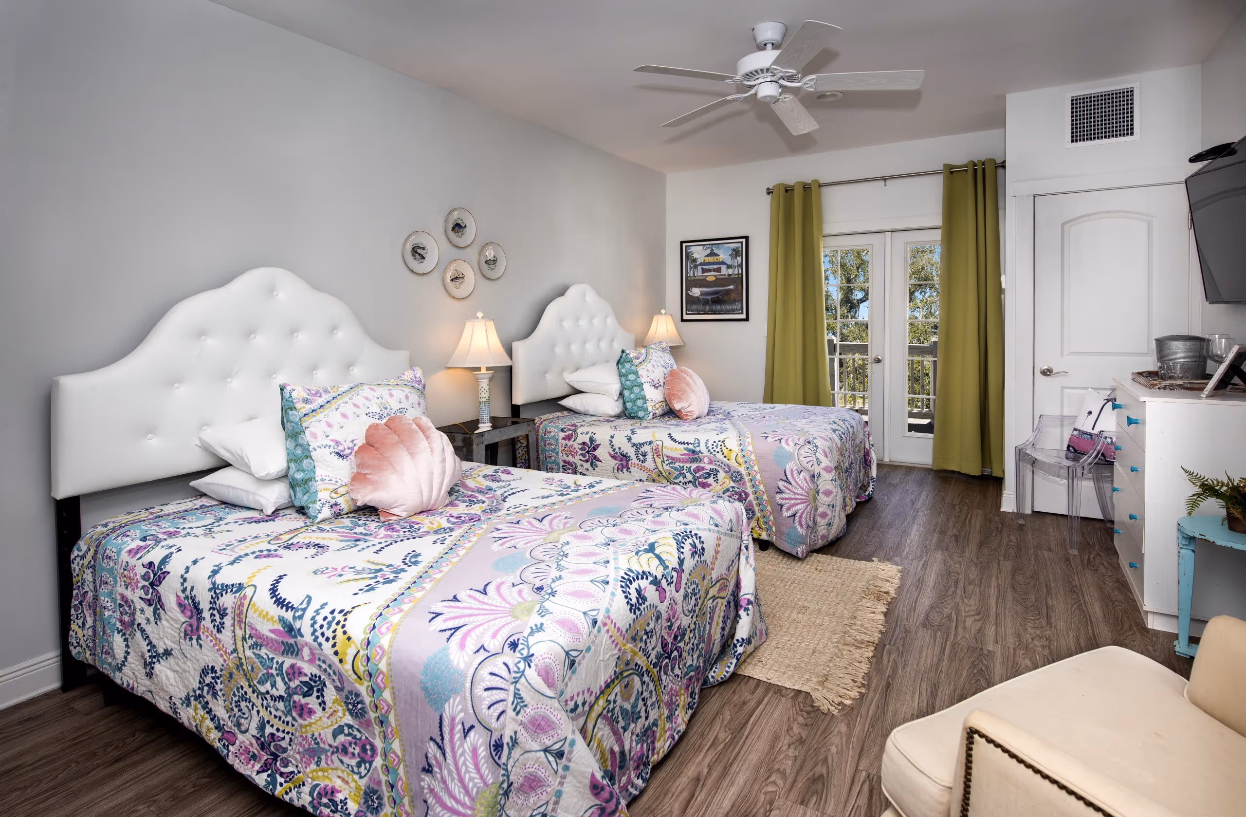 Bright bedroom with two twin beds featuring white tufted headboards, floral bedspreads, a ceiling fan, green curtains, and a TV mounted on the wall.