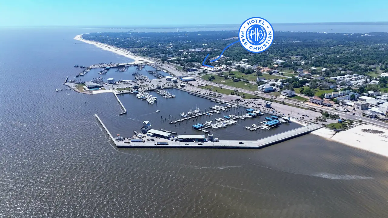 Aerial view of a marina with boats docked and a nearby shoreline with buildings and a hotel logo labeled 'Hotel Pass Christian'.