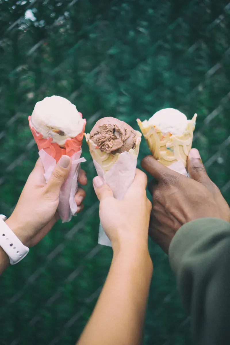 Three hands holding different ice cream cones with green foliage background.