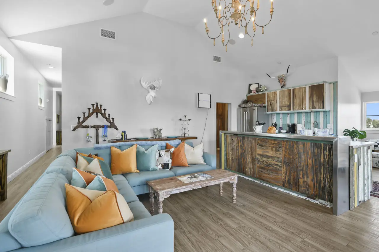 Bright living room with a blue sectional sofa adorned with orange and blue pillows, a rustic wooden coffee table, and a kitchen with distressed wood cabinets and a metal countertop.