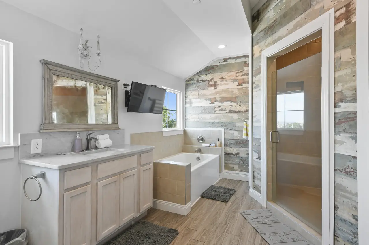 Bright bathroom with a rustic wood-paneled wall, bathtub, glass door shower, wooden vanity with marble countertop, and a wall-mounted TV.