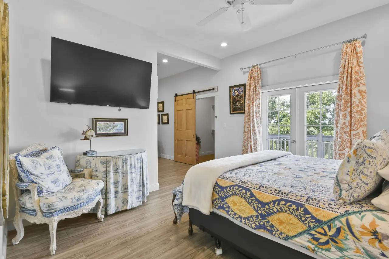 Bright bedroom with patterned bedspread, antique-style chair, wall-mounted TV, and French doors with orange floral curtains.