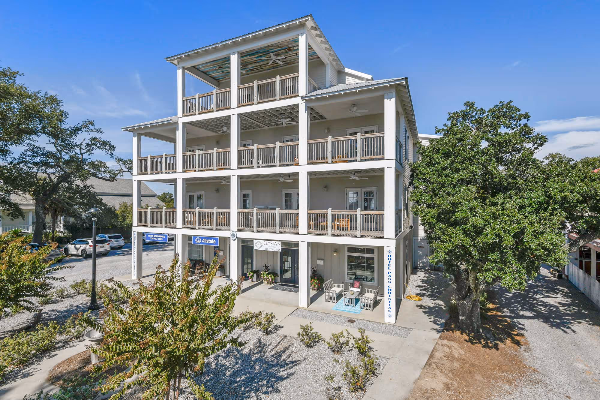 Three-story building with wide balconies, office signs including Allstate, outdoor seating area, and surrounded by trees under a clear blue sky.