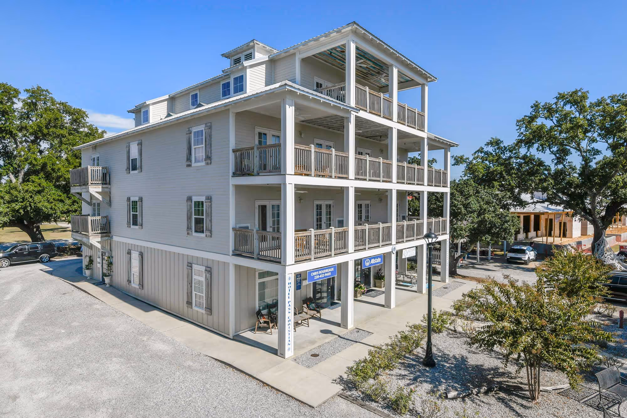 Three-story white building with balconies on each floor, surrounded by trees and parked cars under a clear blue sky.
