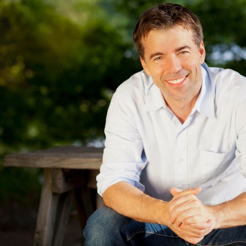 Smiling man in a white shirt sitting outdoors with hands clasped and a blurred green background.