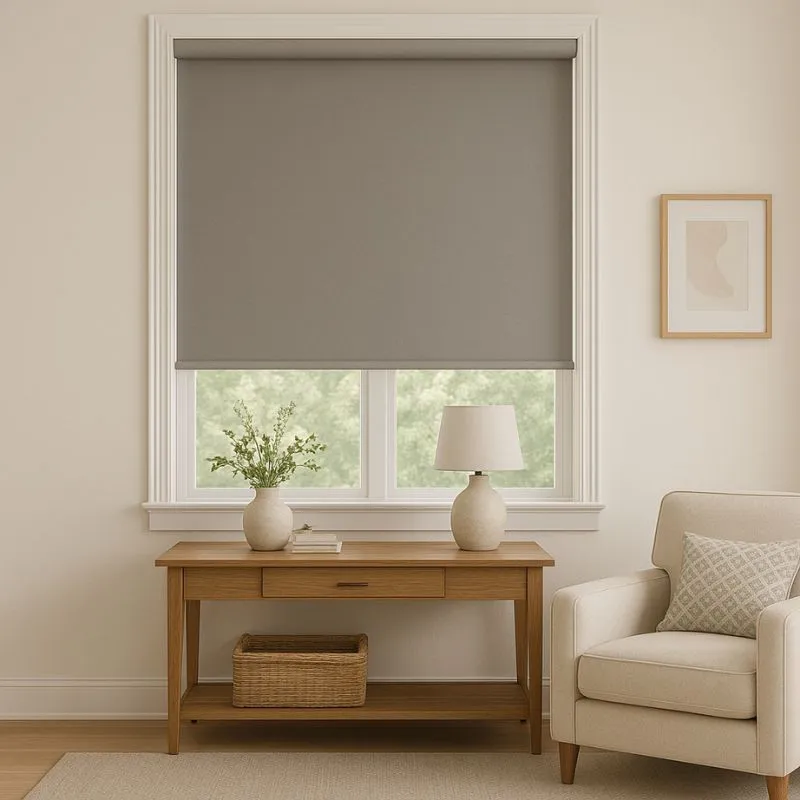 Modern living room with beige roller shade covering a window, wooden console table with a vase, books, lamp, and a wicker basket underneath, beside a beige armchair with patterned pillow.