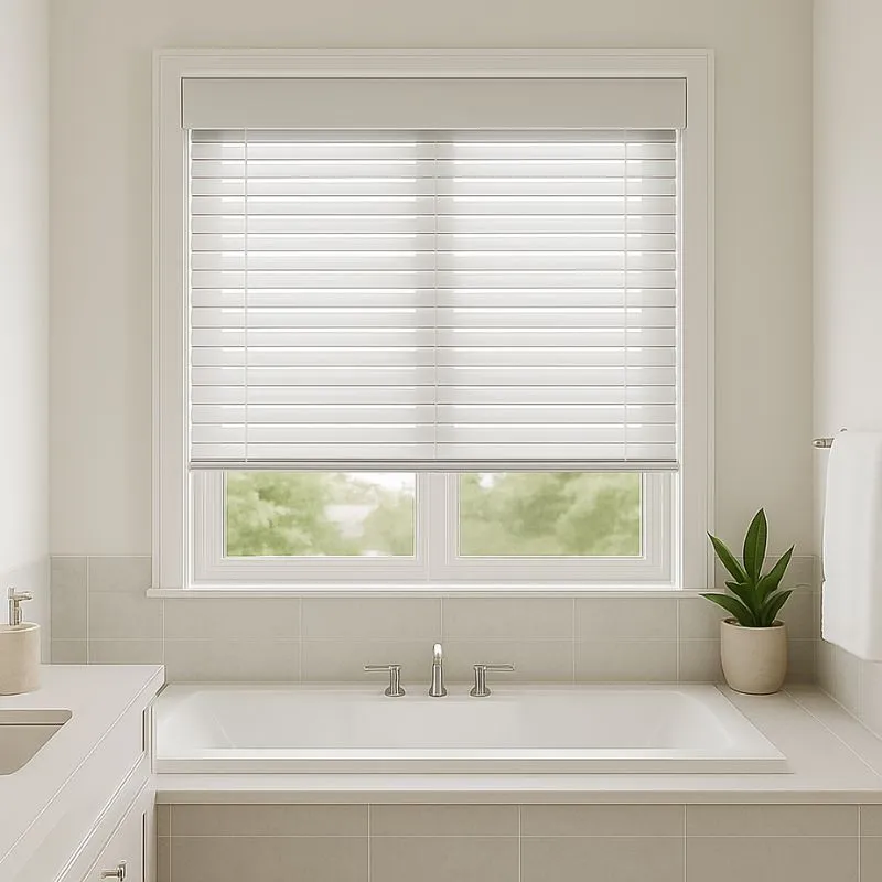 Modern bathroom with white venetian blinds partially covering a double window above a built-in bathtub with silver faucets and a potted plant on the right side.