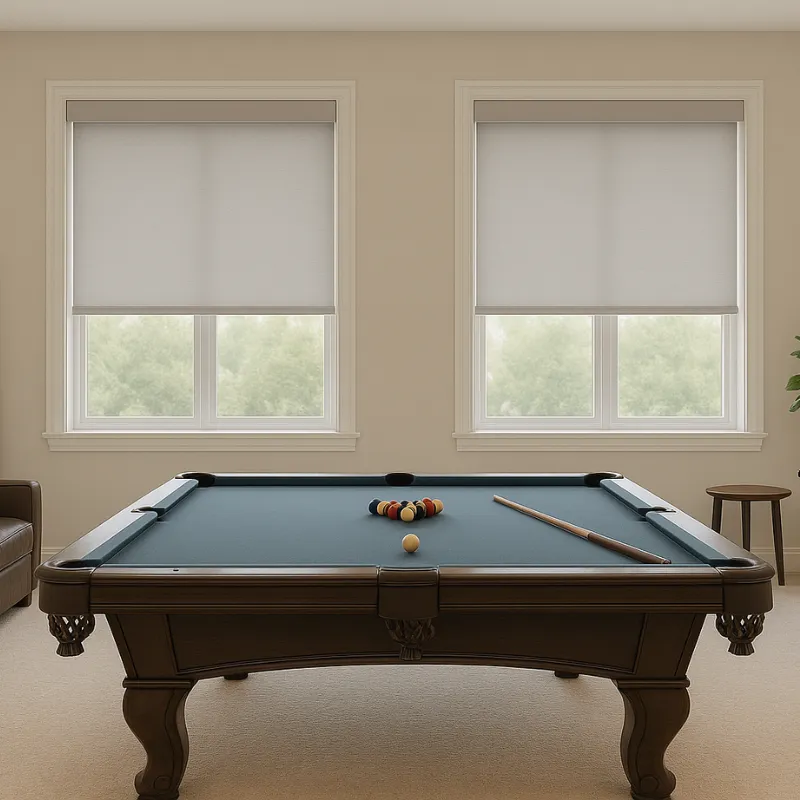 Pool table with billiard balls arranged in a triangle and a cue stick resting on a blue felt surface beneath two windows with white roller blinds.