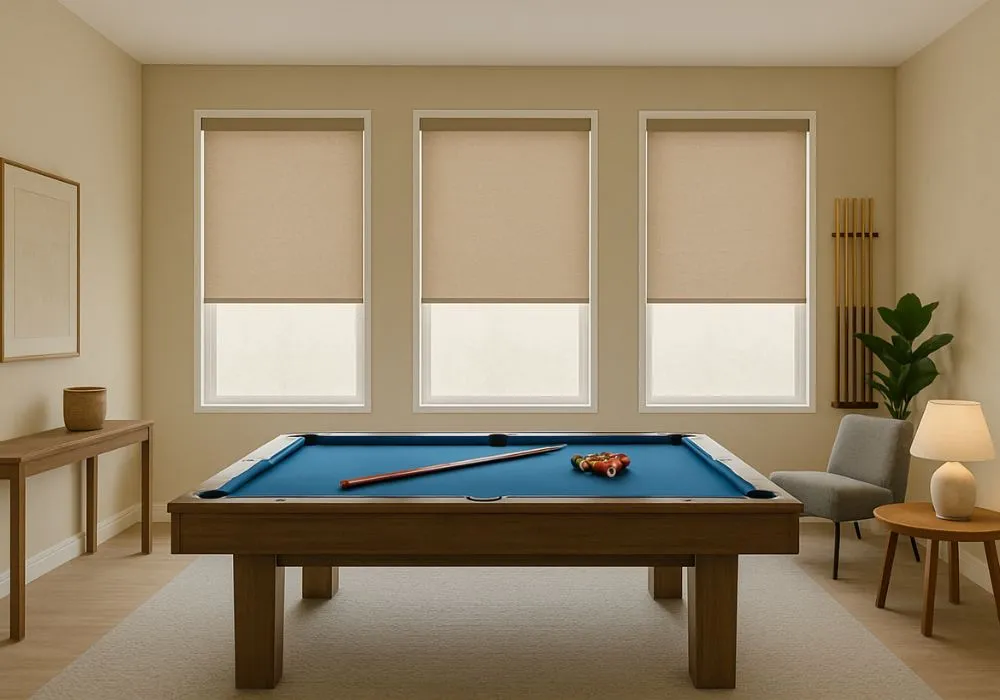 Modern game room with a blue pool table, three beige roller shades covering windows, a gray chair, wooden table, and lamp.